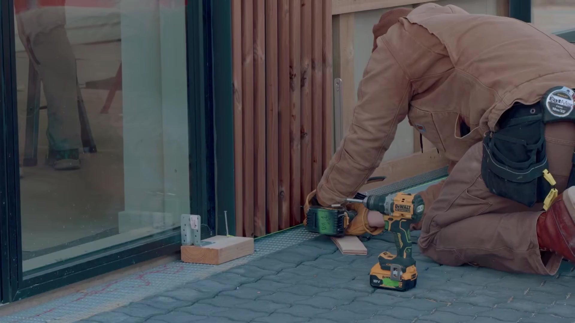 Installer kneeling at the base of a glass facade fixing the bottom profile for vertical Aluwood slat cladding